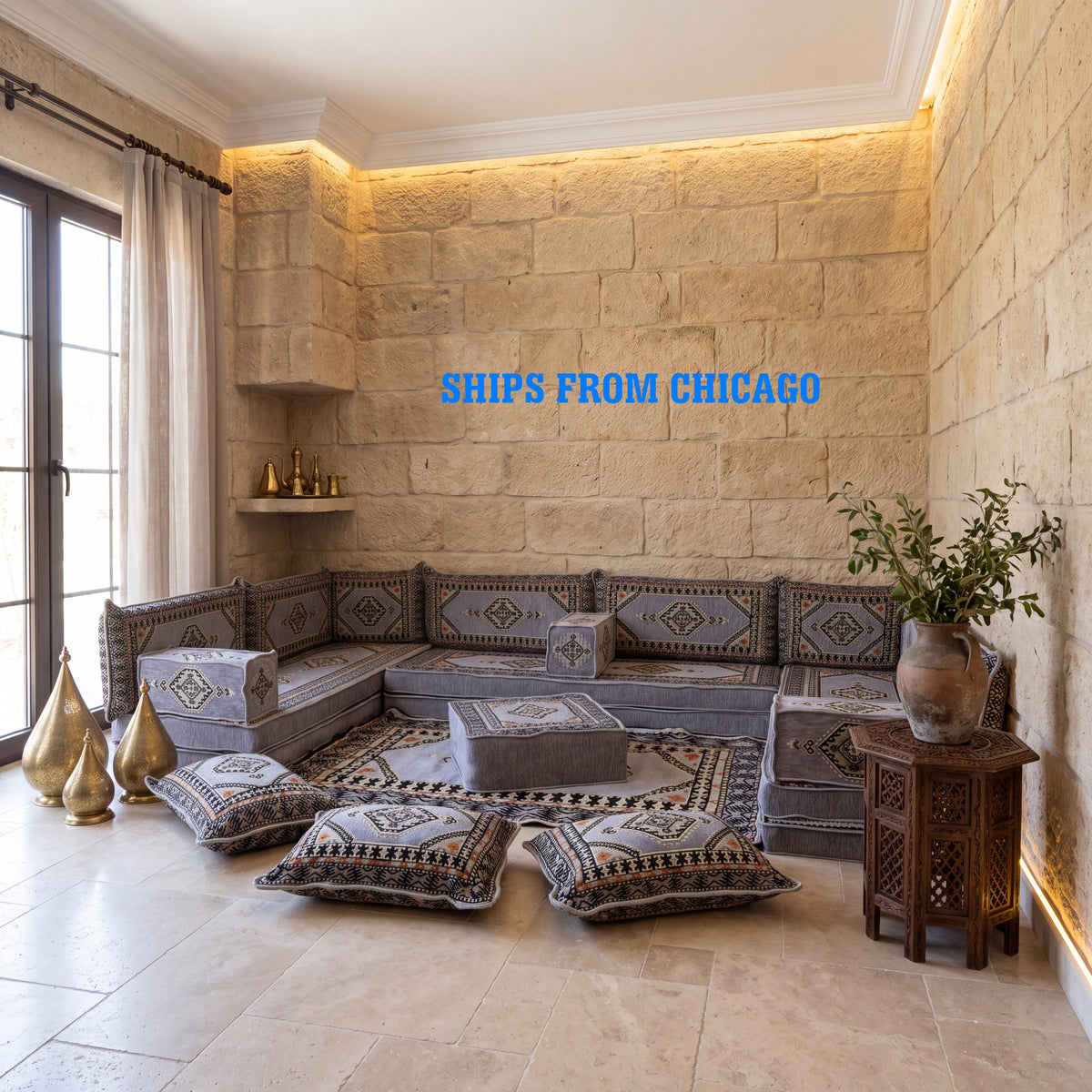 A Arabic-style modular upholstered gray floor seating sofa set arranged in a stone-walled living room. The set includes a low modular sectional sofa, matching armchair, ottoman, and several decorative throw pillows with geometric majlis patterns.
