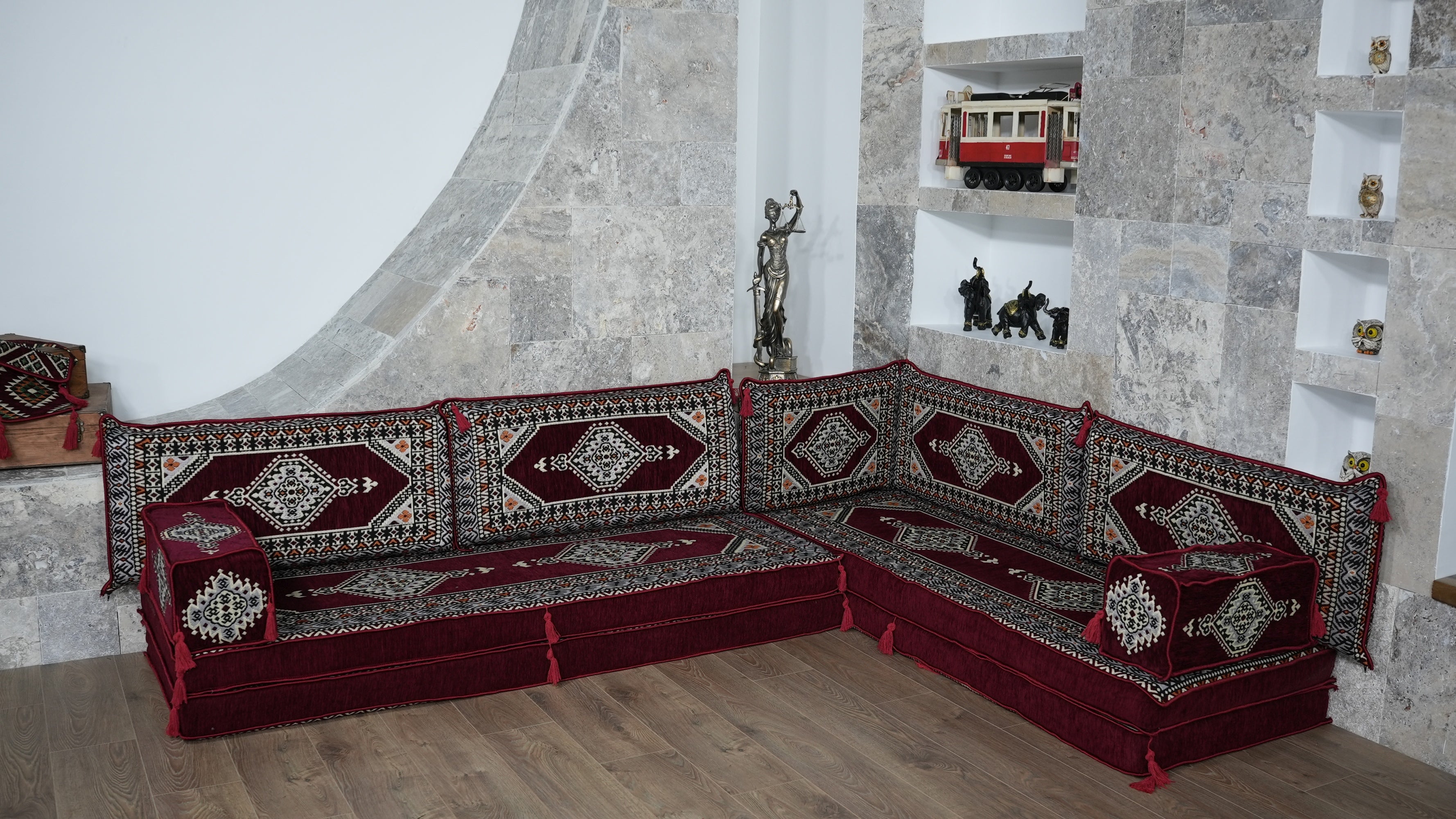 A traditional Turkish Majlis sectional sofa upholstered in deep red, burgundy with intricate white, and gold geometric patterns on the cushions and backrests, set in a stone-walled room.