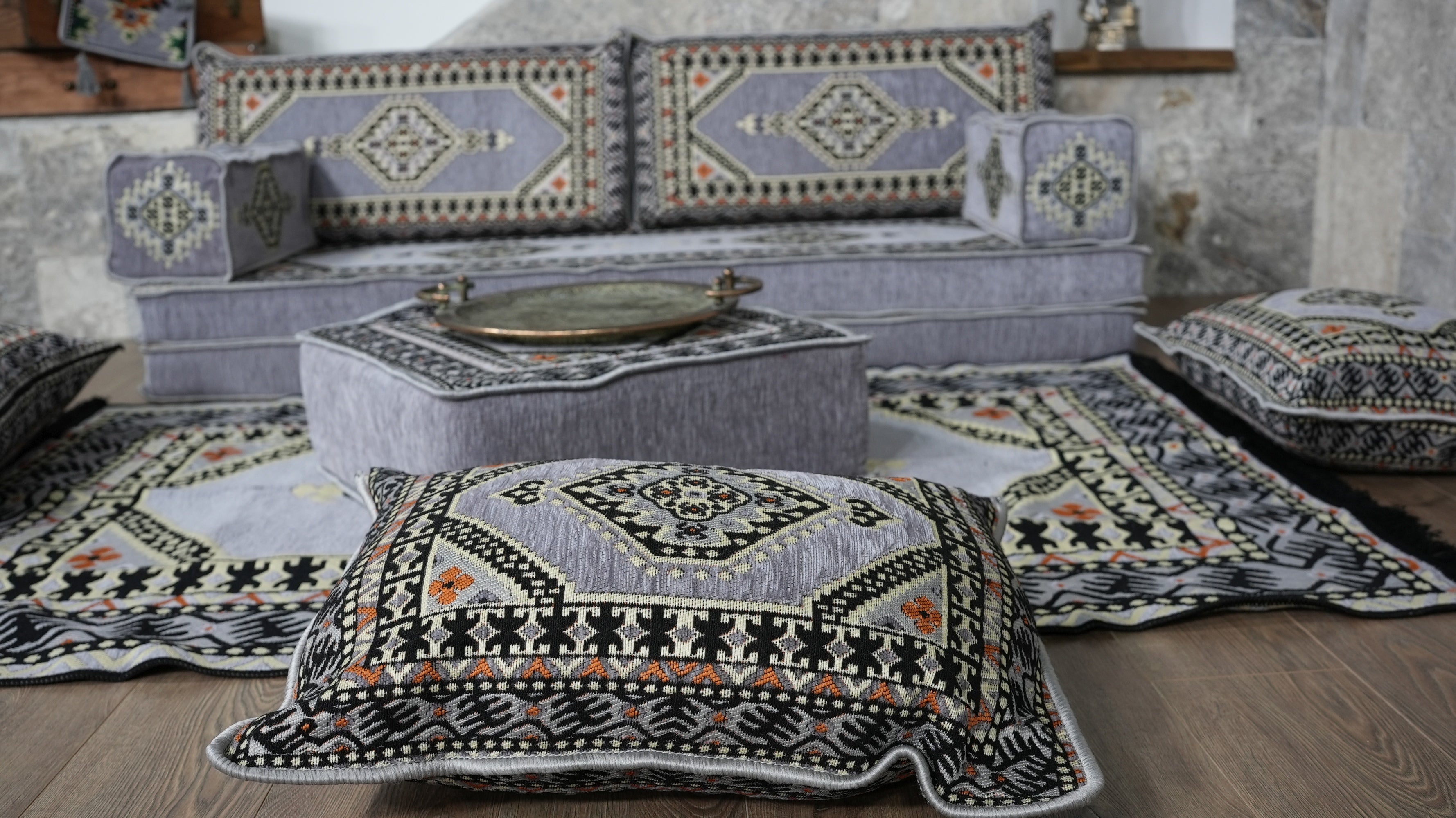 A styled interior scene showing a gray upholstered Arabic Majlis floor sofa with patterned cushions, a matching ottoman, and several decorative throw pillows and a kilim rug.