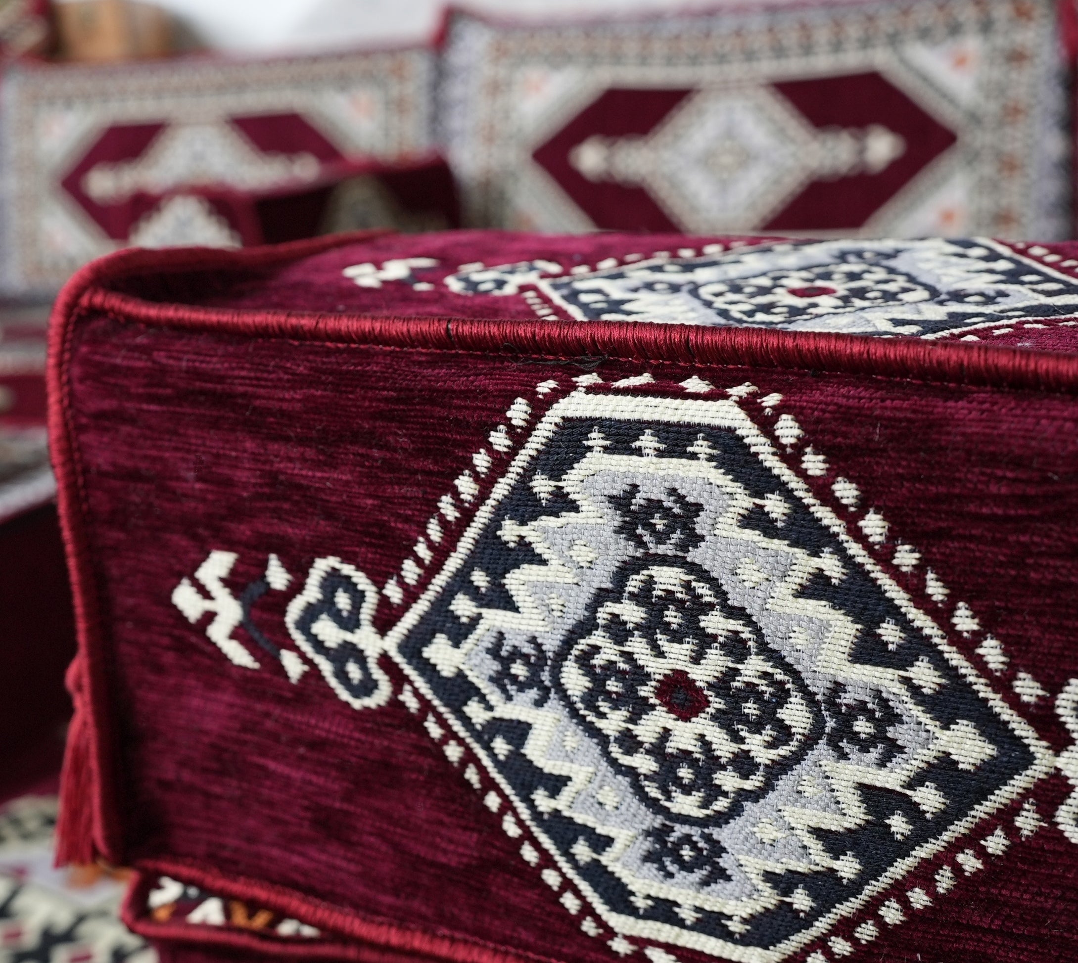 A close-up of a deep burgundy armrest for Arabic majlis sofa with a large geometric medallion pattern in cream and burgundy, with matching patterned textiles blurred in the background.