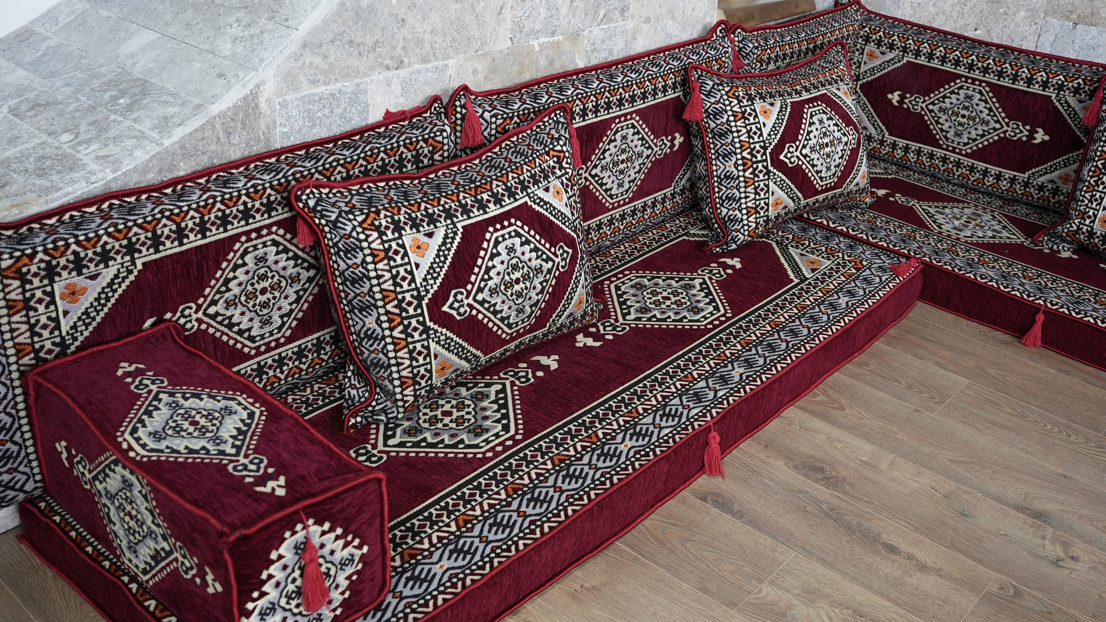 A traditional Ural sofa upholstered in deep red velvet with intricate geometric and floral patterns in beige, black, and white, set against a light stone wall and wooden floor.