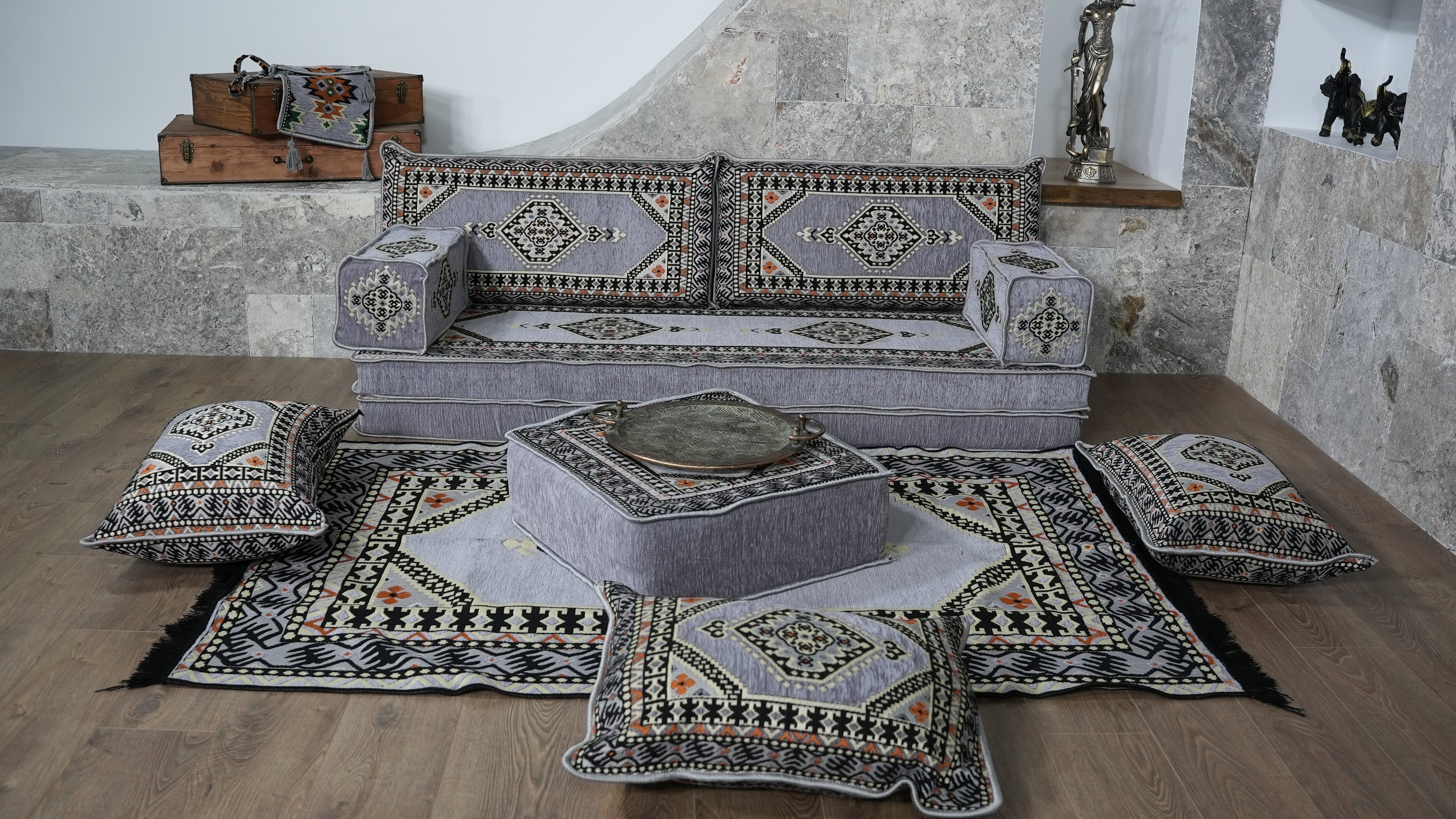 A coordinated set of bohemian-style home decor items including a gray patterned sofa, matching cushions, a low ottoman, and patterned rugs arranged on a wood floor in a stone-walled room.